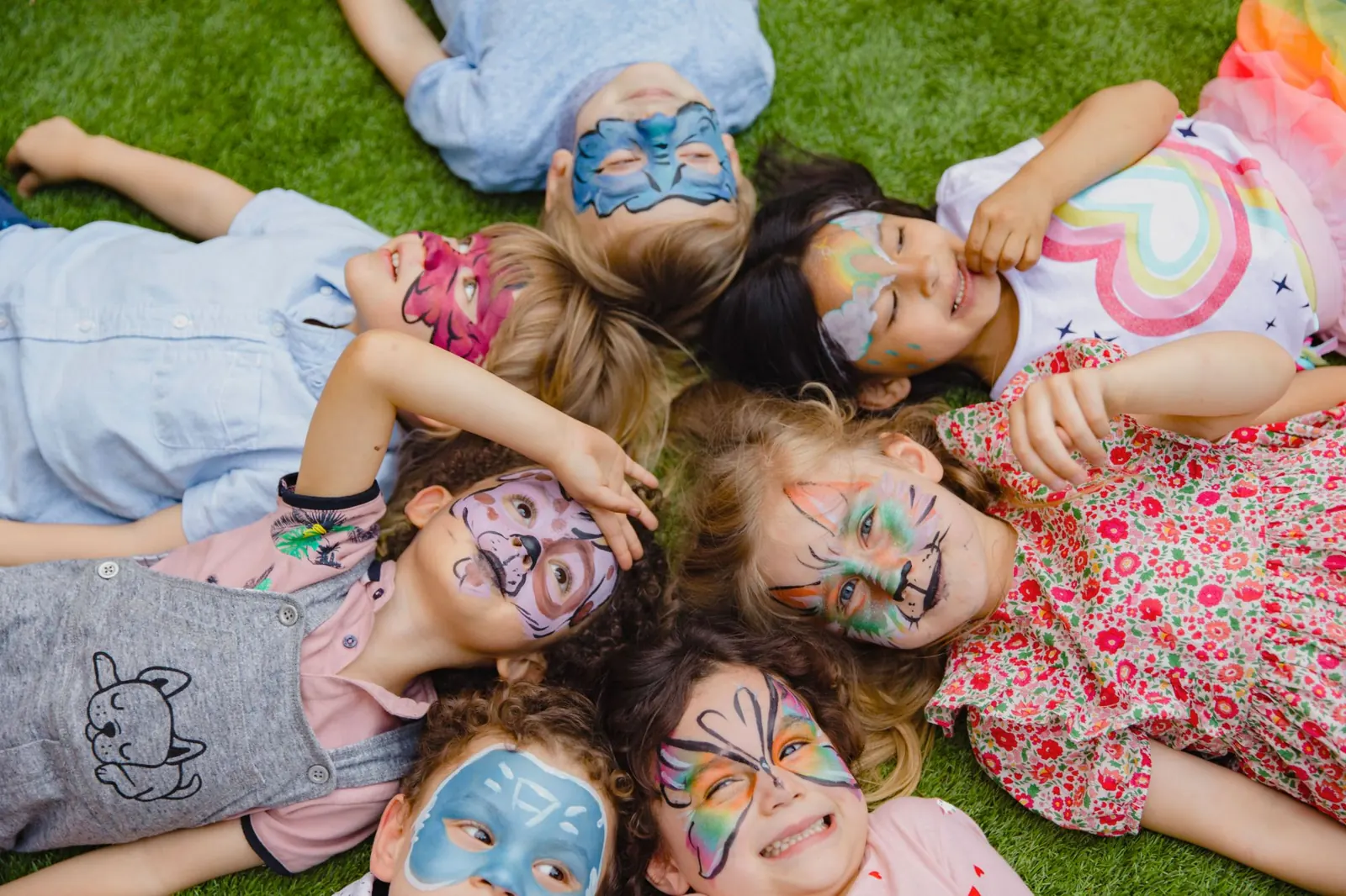 Kinderen met geschminkte gezichten liggen lachend in een kring op het gras